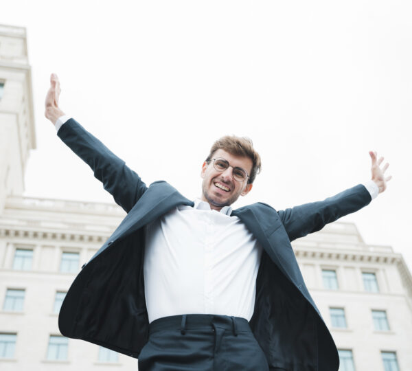 carefree-smiling-young-businessman-standing-front-building-raising-his-arms